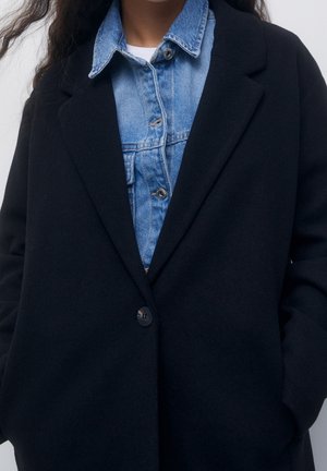 Femme portant des vêtements superposés avec une chemise en denim bleue boutonnée sous un blazer noir à un seul bouton.