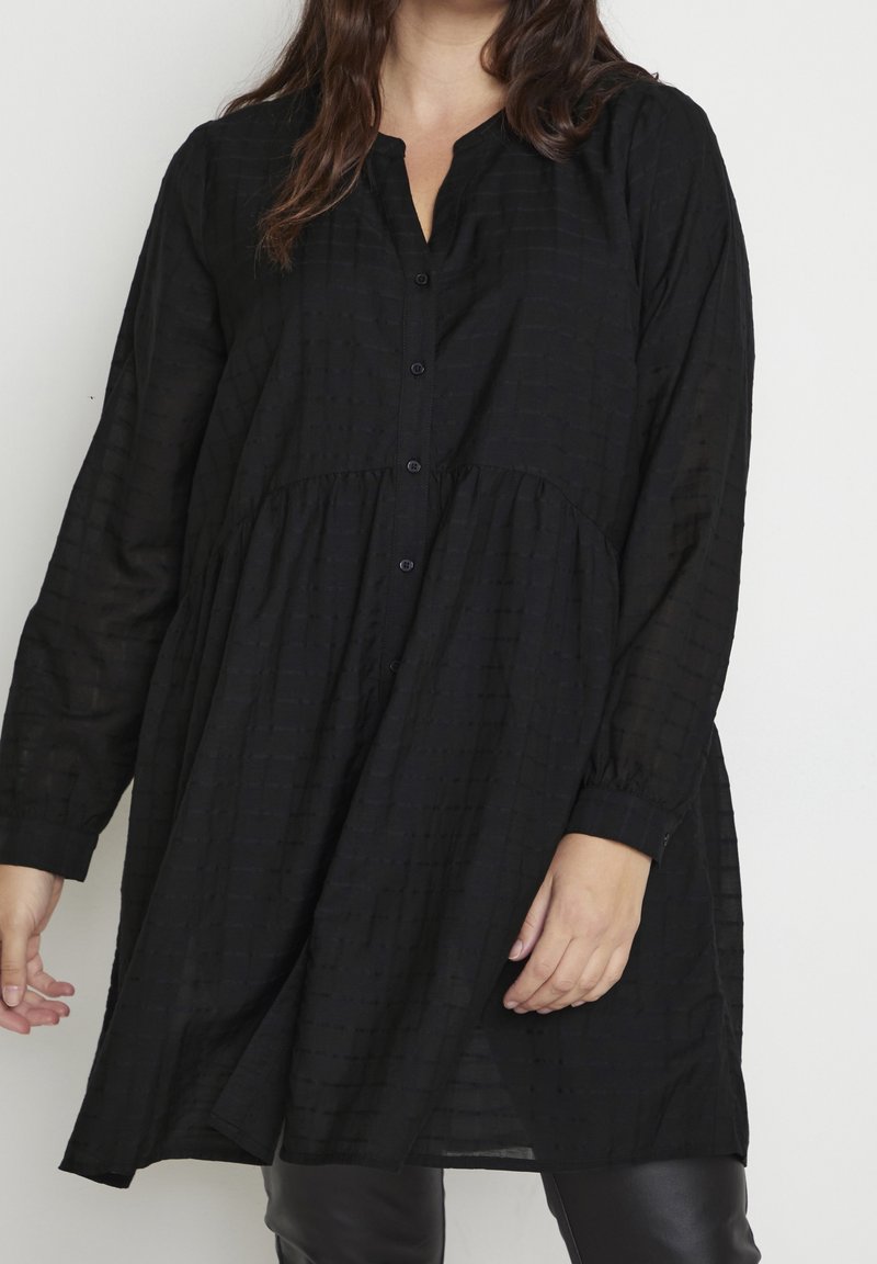 Femme portant une tunique noire ample à manches longues boutonnée sur un pantalon noir, debout devant un fond clair uni.
