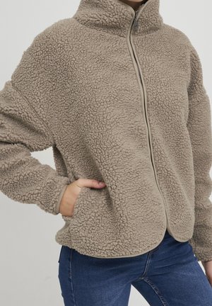 Personne portant une veste polaire beige à col montant zippée, les mains dans les poches et un jean bleu sur un fond uni.