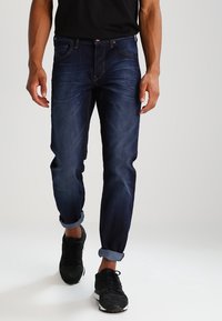 Jeans de mezclilla azul oscuro con un corte ajustado, leve desvanecimiento, puños enrollados y diseño de cinco bolsillos. Combinados con zapatillas negras.
