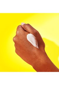 Hand with brown skin shown against a yellow background, featuring a thick, white cream applied on the back. Smooth texture visible.