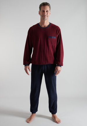 Man standing barefoot wearing a burgundy long-sleeve top with a chest pocket and dark blue pants against a plain light background.