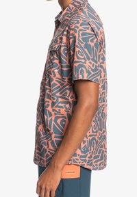 Koszula z krótkim rękawem z geometrycznym wzorem w kolorach różowym i niebieskim, wykonana z teksturowanej tkaniny, z dwoma kieszeniami na piersi oraz metką z nazwą marki przy dolnym brzegu.