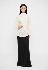 Top de manga longa com padrão floral branco, combinado com uma saia preta até ao chão. A modelo segura uma clutch creme com detalhe de fecho metálico.