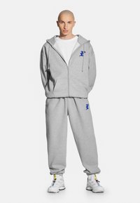 Szara bluza z kapturem na zamek błyskawiczny z niebieskim logo, w zestawie ze spodniami dresowymi w tym samym kolorze. Posiada kaptur ze sznurkiem, luźny krój i elastyczne mankiety.