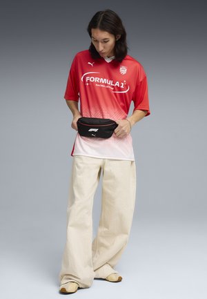Personne en t-shirt rouge Formula 1 Racing League, pantalon beige à jambes larges et baskets, ajustant une banane noire Puma sur un fond gris.