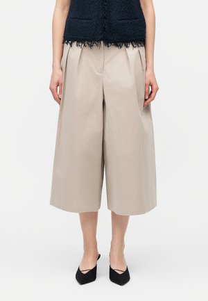 Moteris dėvi smėlio spalvos plačių kojų kelnės su priekinėmis klostėmis, derinamos su juodais smailiais sandalais su dirželiais ir juodu tekstūruotu viršutiniu drabužiu.