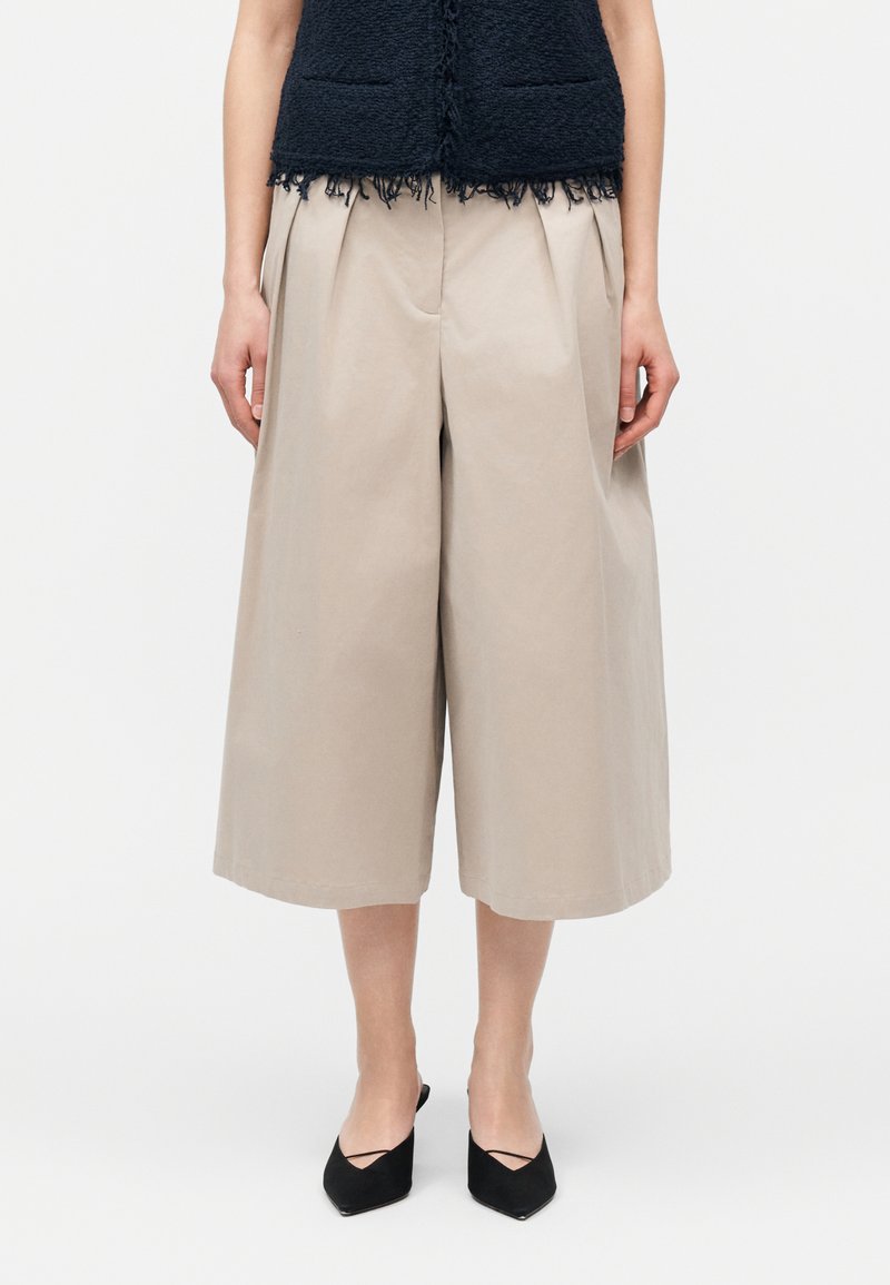 Žena oblečená v béžových širokých nohaviciach culotte s prednými záhybmi, doplnená čiernymi lodičkami so špicatou špičkou a čiernym textúrovaným topom.