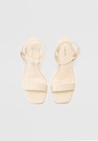 Beige stroppesandaler med flat såle, med en tynn ankelstropp og en bredere frontstropp. Glatt tekstur og minimalistisk design.