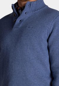 Marineblauwe gebreide trui met hoge kraag, drieknoopsluiting, geribbelde halslijn en gestructureerde stof. Subtiele logodetail op de borst.