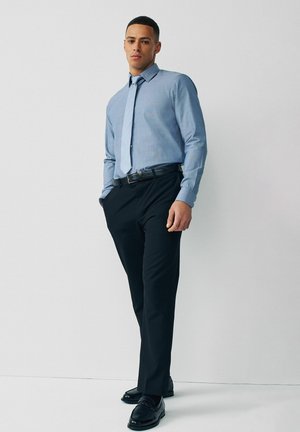 Noor mees, kes kannab helesinist triiksärki, sobivat lipsu, musti pükse ja musti kingi, seisab valge seina taustal ühe käega taskus.