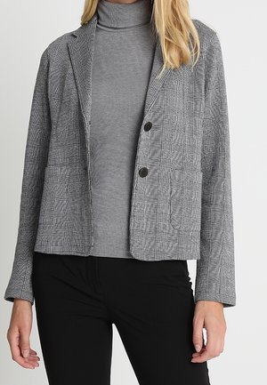 Donna che indossa un blazer grigio a quadri con due bottoni sopra un dolcevita grigio e pantaloni neri contro uno sfondo semplice.