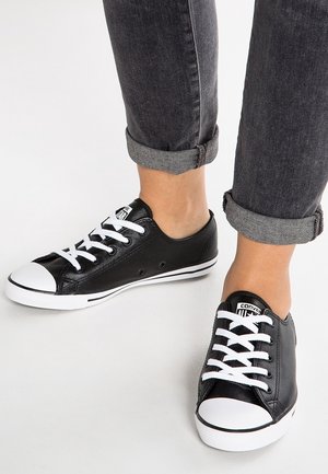 Tummanharmaat rullatut farkut, joissa mustat matalavartiset tennarit, joissa on valkoiset nauhat, kärjet ja pohjat, vaaleanharmaalla lattialla.