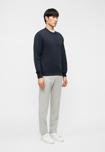 Μπλε βαμβακερό φούτερ με στρογγυλή λαι μόκοψη, ριμπ μανσέτες και τελείωμα; σε συνδυασμό με ανοιχτό γκρι παντελόνια και λευκά αθλητικά παπούτσια. Διακριτική λεπτομέρεια λογότυπου στο στήθος.