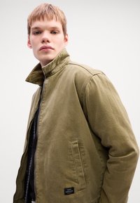 Young person with short hair wearing an olive green zip-up jacket with a raised collar against a plain white background.