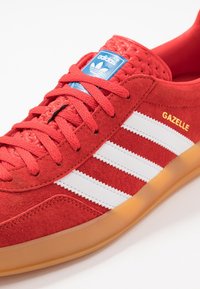 Röd mockaskor med tre vita läderstrimmor och "Gazelle"-logotyp. Har texturerad röd foder, rosa snören och en genomskinlig gummsula.