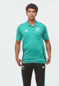 Młody mężczyzna ubrany w turkusową koszulkę polo i czarne sportowe spodnie z logo Adidas oraz New Zealand All Blacks, stojący na jednolitym tle.