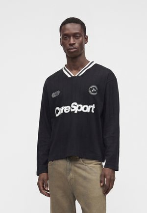Mand iført en sort langærmet sportstrøje med hvide kanter og logoer, kombineret med beige bukser, stående mod en ensfarvet hvid baggrund.