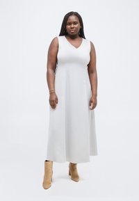 Γυναίκα με μακριά πλεξούδα, ντυμένη με αμάνικο ανοιχτό γκρι φόρεμα και καφέ μποτάκια αστραγάλου, στέκεται σε απλό λευκό φόντο.