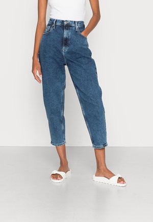 Personne portant un jean bleu taille haute, fuselé, un top blanc sans manches et des sandales blanches, debout devant un fond uni.