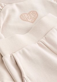 Világos bézs pulóver bordázott nyakkal és mandzsettával, szív alakú logófolttal, amely texturált mintázatot tartalmaz. Puha anyag.
