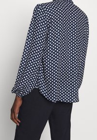 Blusa blu navy con motivo a cuore bianco, dotata di maniche lunghe con polsini arricciati, chiusura a bottoni sul retro e una vestibilità rilassata.