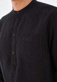 Homme portant une chemise noire à manches longues avec un col mandarin, une patte de boutonnage et une poche poitrine visible sur le côté gauche.