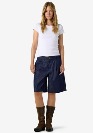 Jeune femme debout vêtue d'une chemise blanche à manches courtes, d’un short bleu foncé jusqu’aux genoux et de bottes marron mi-mollet sur fond blanc.