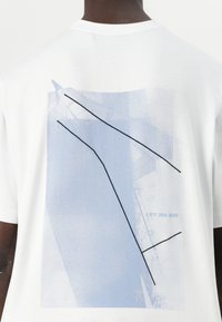 White cotton t-shirt featuring a blue graphic print with abstract lines and shapes, including black accents and the text "J K11 200 0HB."