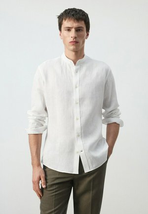 Hombre vistiendo una camisa blanca de manga larga con cuello mandarín y mangas remangadas, combinada con pantalones oscuros, de pie contra un fondo liso.