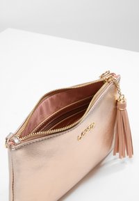 Bolso de mano en oro rosa con cremallera y borla doradas, parcialmente abierto para mostrar los compartimentos interiores y el forro de tela suave.