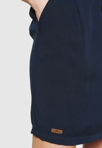 Marinblå kjol i mjukt tyg, med sidfickor, veckdetalj och en liten brun etikett vid fållen.