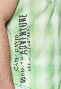 Camisa de manga corta verde claro con un patrón de cuadrícula, con texto negro que dice "CAMP DAVID AVENTURE GREEN DEPARTMENT" en el lateral.