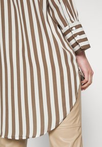 Main portant un pantalon beige et une chemise longue à rayures verticales marron et blanches, avec des poignets boutonnés et une fente latérale, sur un fond uni.