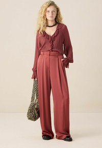 Vrouw met golvend blond haar, gekleed in een roestkleurige blouse met ruches en een brede broek, die een tas met luipaardprint vasthoudt, staande tegen een beige achtergrond.