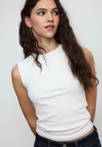 Top bianco senza maniche con una texture liscia e scollo rotondo, abbinato a jeans di denim scuro. I capelli della modella sono mossi e di lunghezza fino alle spalle.