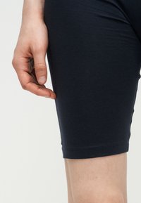 Prawa ręka swobodnie opuszczona obok uda, nosząca czarne obcisłe szorty na jasnym tle.