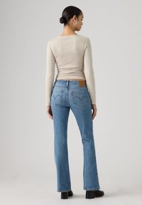 Γυναίκα με σκούρα μαλλιά δεμένα κότσο, που φοράει μπεζ μακρυμάνικη μπλούζα, μπλε τζιν με ίσια κορδόνια και μαύρα παπούτσια με τακούνι, στέκεται με την πλάτη προς το γκρι φόντο.