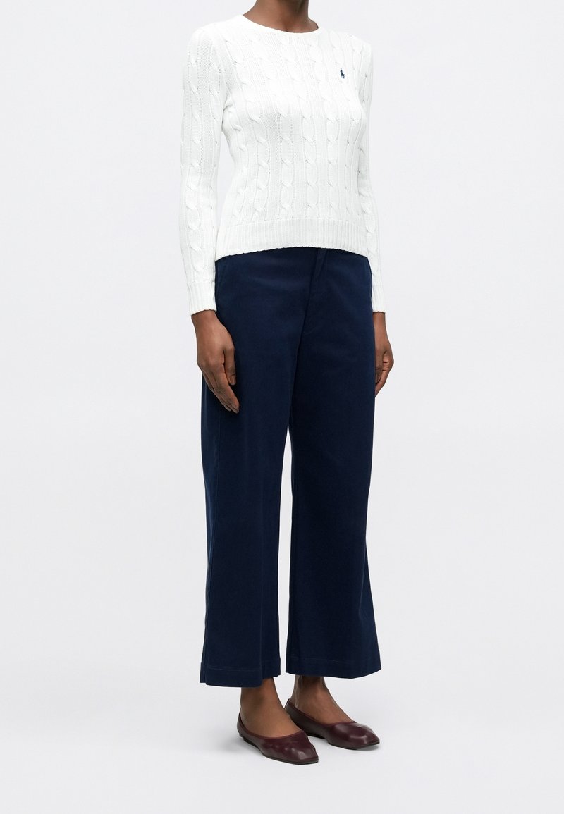 Femme portant un pull en maille torsadée blanc, un pantalon large bleu marine et des chaussures plates rouge foncé, debout devant un fond uni.