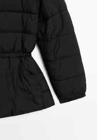 Dettaglio di giacca a vento nera con design trapuntato, polsini elasticizzati, vita cinchata e tasche laterali. Texture liscia e lucida.