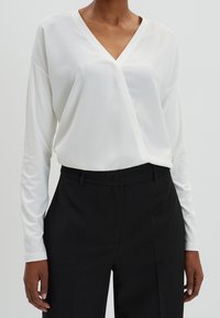 Top bianco a maniche lunghe avvolgente con scollatura a V, tessuto testurizzato e vestibilità comoda, abbinato a pantaloni neri su misura. Design minimalista.