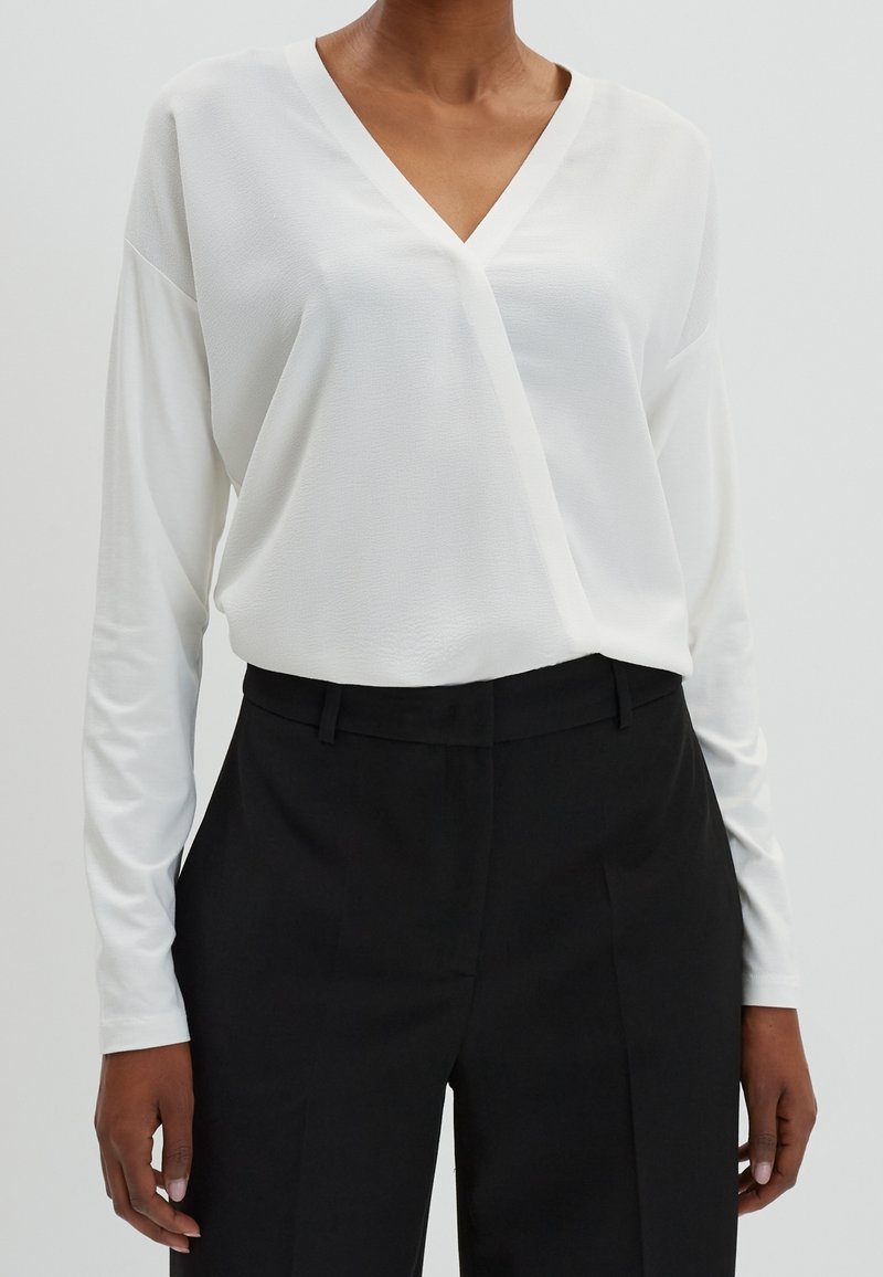 Top bianco a maniche lunghe avvolgente con scollatura a V, tessuto testurizzato e vestibilità comoda, abbinato a pantaloni neri su misura. Design minimalista.