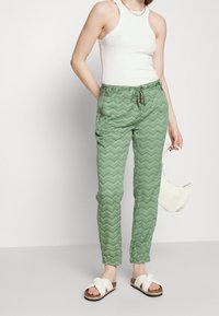 Woman wearing white sleeveless top, green patterned pants, white sandals, holding small white handbag against plain background.