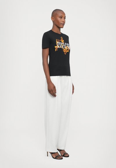 T-shirt en coton noir avec un imprimé graphique présentant des flammes et du texte. Assorti avec un pantalon taille haute blanc à rayures et des sandales à talons noires.