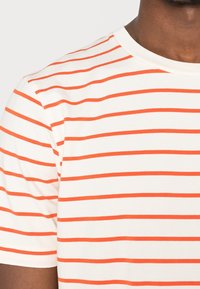 Hombre vestido con una camiseta blanca de manga corta con rayas naranjas horizontales, recortada para mostrar la parte superior del torso y el cuello.