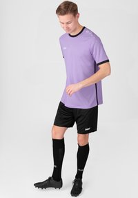 Paarse sporttrui met zwarte accenten, gecombineerd met zwarte shorts en knielange zwarte sokken. De speler draagt zwarte voetbalschoenen.
