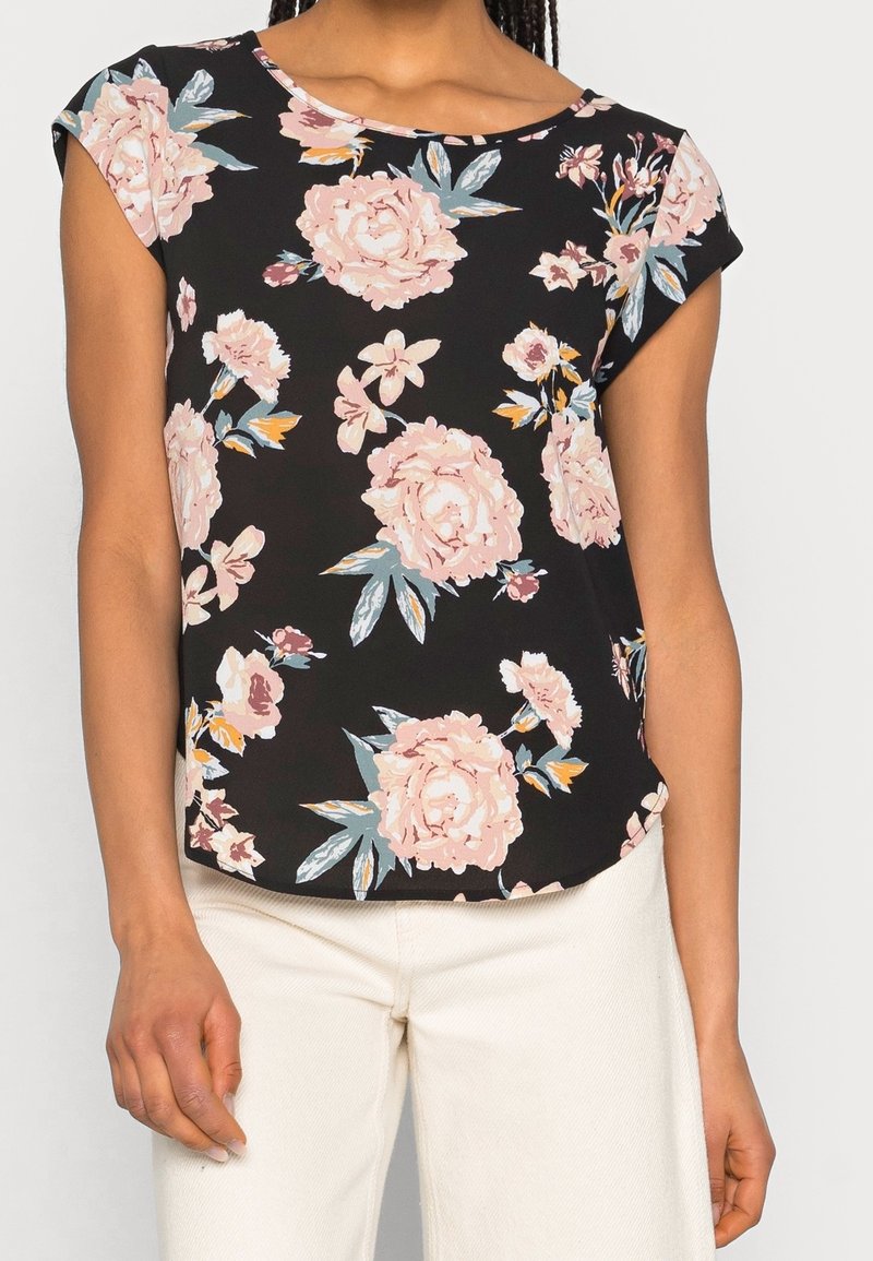 Femme portant un chemisier noir à manches courtes avec un imprimé floral rose et blanc, associé à un pantalon clair, debout devant un fond uni.