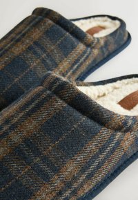 Uldslippers med et marineblåt plaidmønster, der har en blød cremefarvet foret og en tekstureret overflade. Brune læderdetaljer indeni.