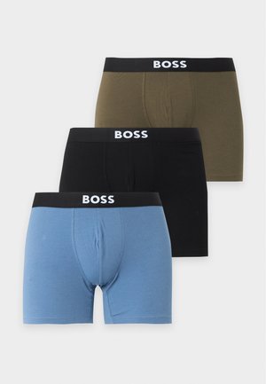 Homme aux cheveux foncés portant un boxer bleu avec une ceinture noire indiquant BOSS, debout pieds nus sur un fond blanc.