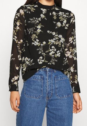 Zwarte doorzichtige blouse met lange mouwen en witte bloemenprint, gecombineerd met hooggetailleerde blauwe denim jeans met voorzakken.
